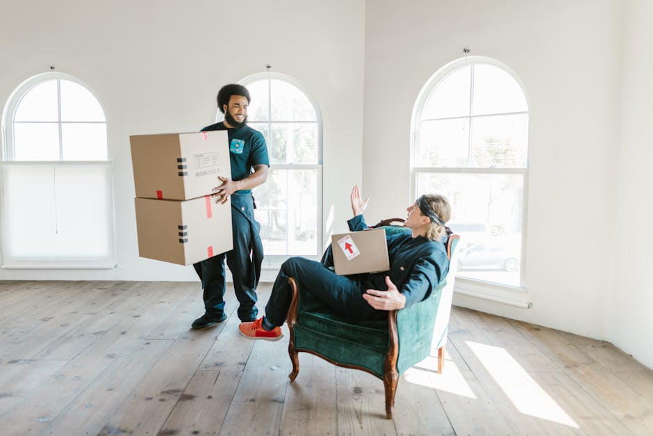 Two women are standing inside a house surrounded by stacked cardboard moving boxes, some sealed with red packing tape. One woman, dressed in a white top and beige pants, holds a medium-sized box with